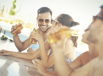 Group of mid 20's people having fun at beach bar. There are two guys and two girls on vacation together. One of the guys is in focus,facing camera and smiling. Each person is having beer and wearing sunglasses.