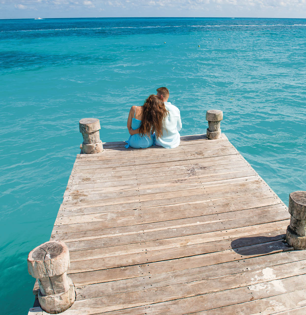 Couple siting at the wooden pier, hugging and looking at the turquoise water of caribbean sea in Cancun. Drone view picture.