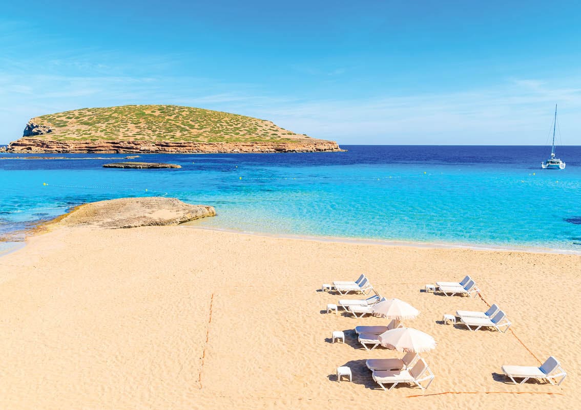 View of beautiful Cala Comte beach with sunbeds and umbrellas - famous for its azure crystal clear shallow sea water, Ibiza island, Spain