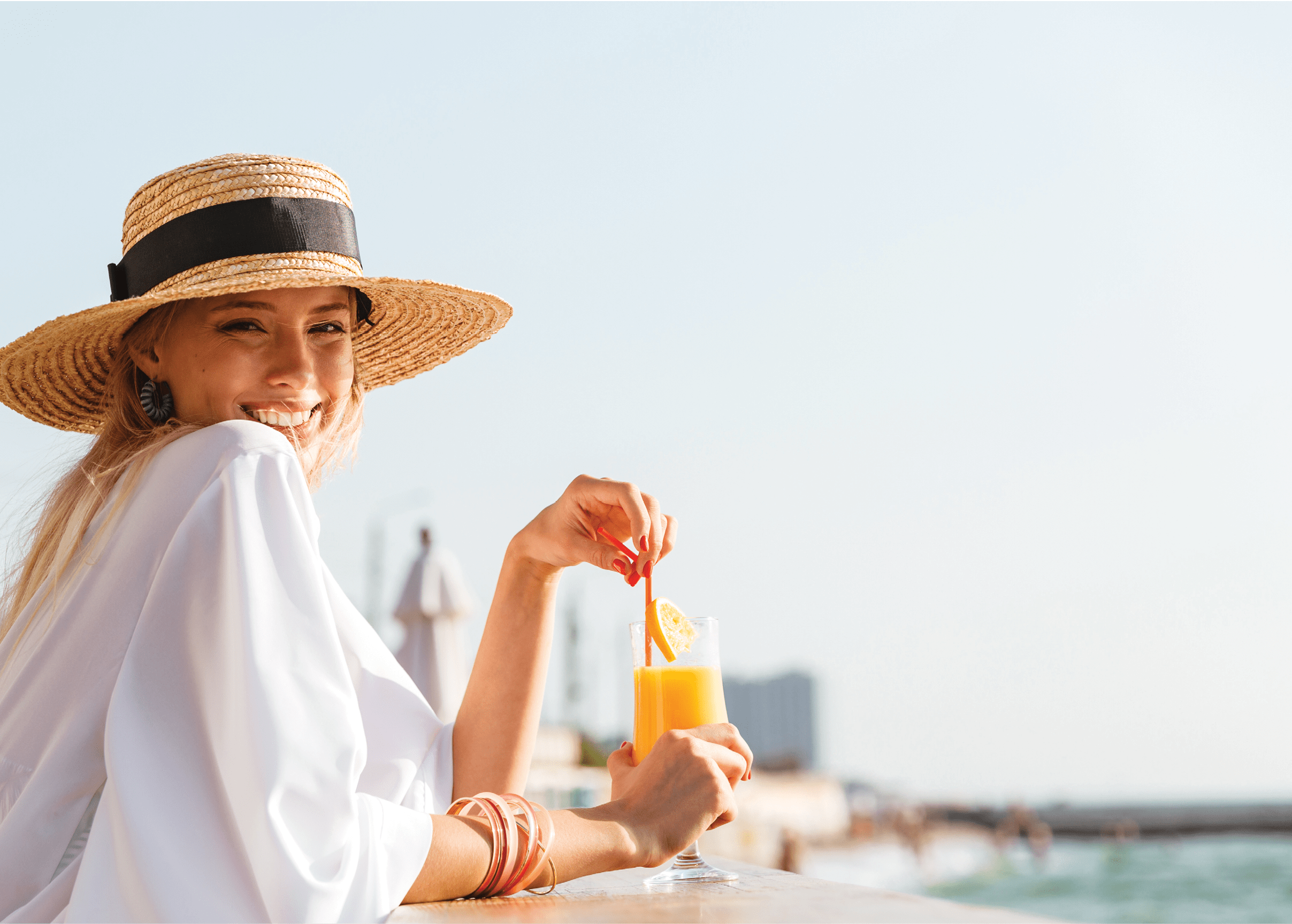 Delighted young girl in summer hat and swimwear resting at the sunny beach, sitting at a cafe with a cocktail