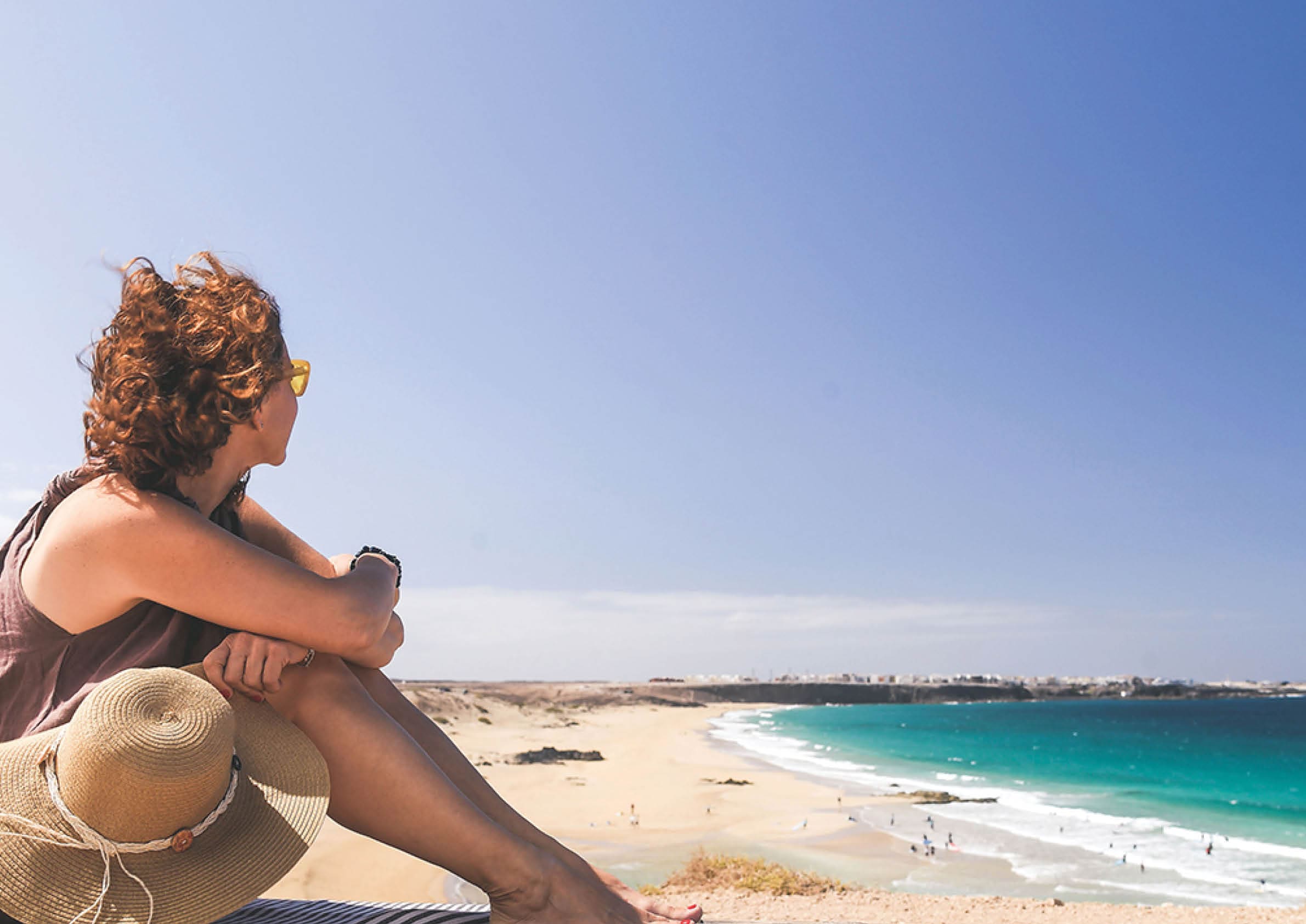 Female tourist looking the tropical panorama in a windy day  Amazing ocean beach in Fuerteventura Spain  Beautiful girl relaxing on a wooden bench  Travel, vacation, happiness and carefree concept