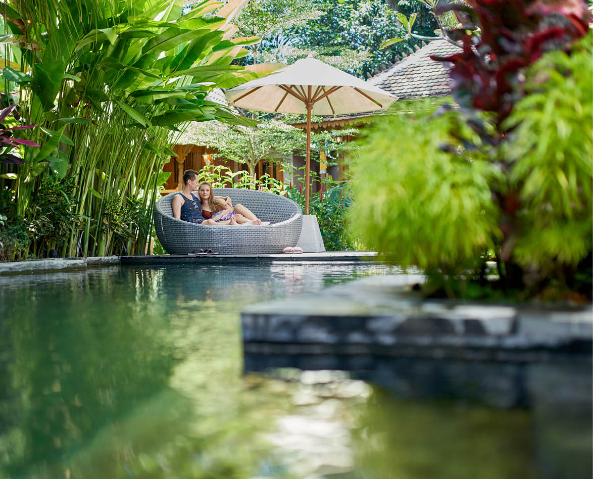 Natural shot of caucasian man and woman lounging outside together by a luxurious pool, during their romantic vacation at an exotic plant-filled villa in Bali offering spa and wellness services