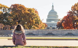Young woman in coat sitting looking at view of United States Congress Capitol building, golden orange yellow foliage autumn fall trees on street during sunny day in Washington DC