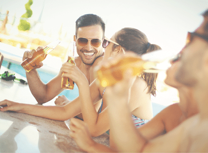 Group of mid 20's people having fun at beach bar. There are two guys and two girls on vacation together. One of the guys is in focus,facing camera and smiling. Each person is having beer and wearing sunglasses.