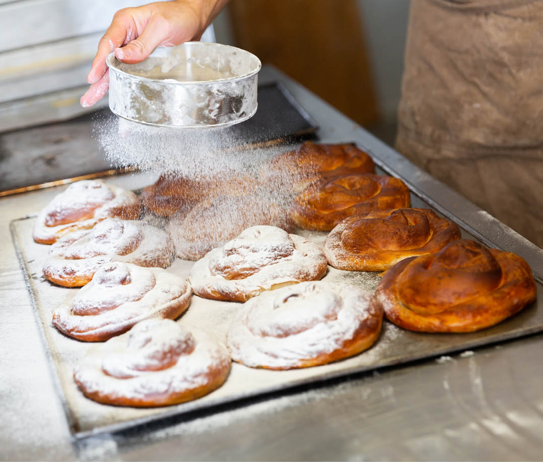 Baker hands dusting freshly baked ensaimadas with powdered sugar in bakery. Baking process of traditional Spanish pastries