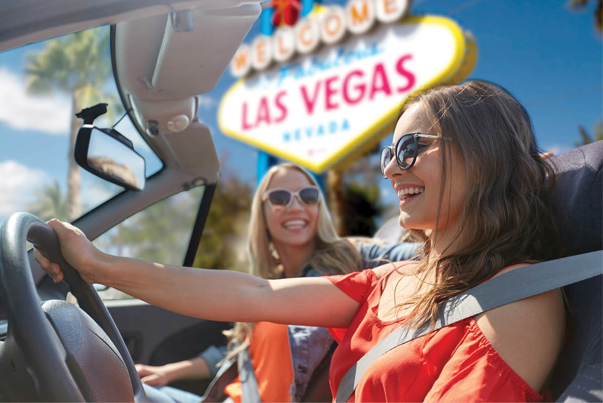 summer holidays, road trip and travel concept - happy young women driving in convertible car and laughing over welcome to fabulous las vegas sign background