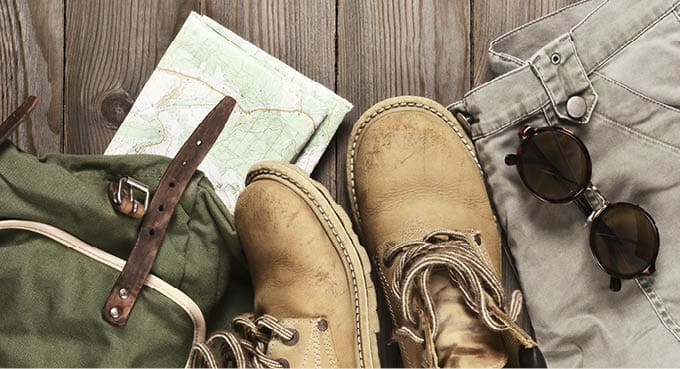 Travel accessories set on wooden background: old hiking leather boots, pants, backpack, map and sunglasses. Top view point.