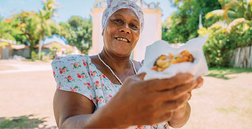 Happy Brazilian woman dressed in the traditional Bahian costume of the Umbanda religion, offering acaraj © - typical Bahia food - in the historic center of Porto Seguro in the background