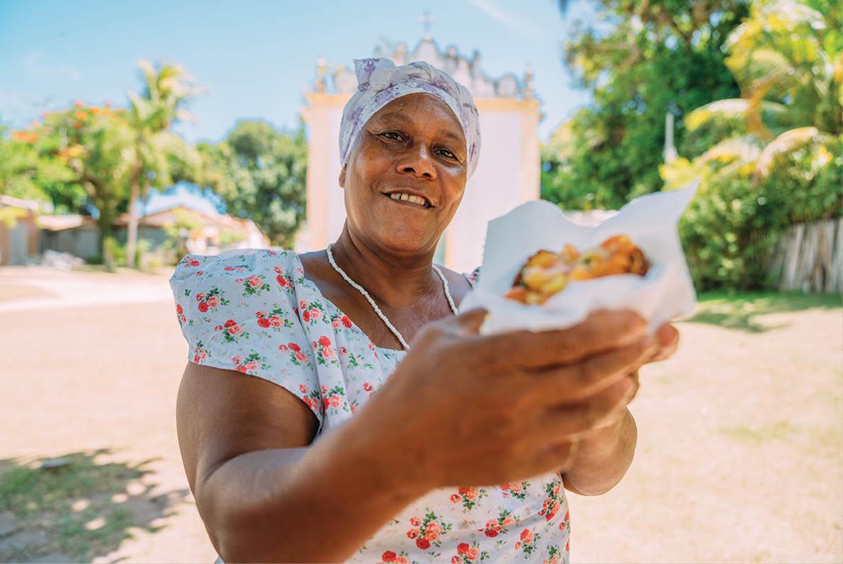 Happy Brazilian woman dressed in the traditional Bahian costume of the Umbanda religion, offering acaraj © - typical Bahia food - in the historic center of Porto Seguro in the background