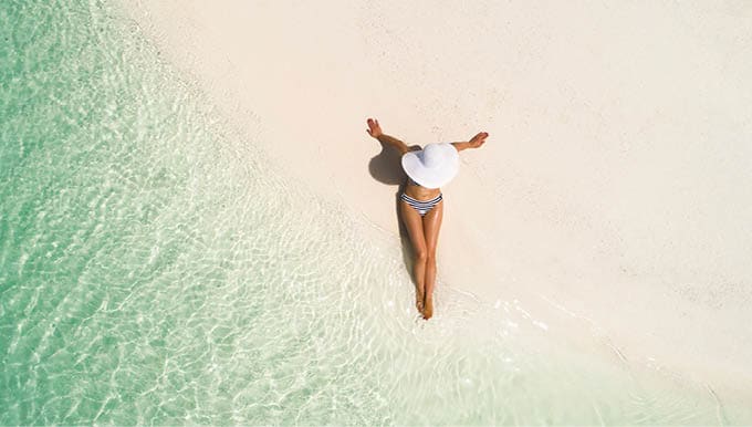 Summer holiday fashion concept - tanning girl wearing sun hat at the beach on a white sand shot from above.Top view from drone. Aerial view of slim woman sunbathing lying on a beach in Maldives.
