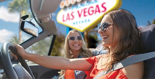 summer holidays, road trip and travel concept - happy young women driving in convertible car and laughing over welcome to fabulous las vegas sign background