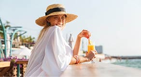 Delighted young girl in summer hat and swimwear resting at the sunny beach, sitting at a cafe with a cocktail