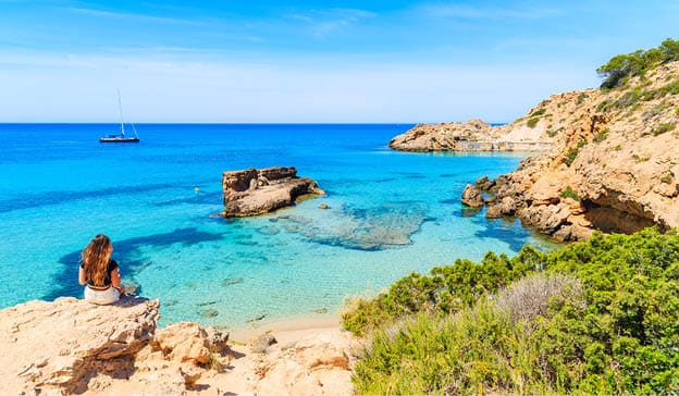 Unidentified young woman sitting on a rock and looking at beautiful Cala Tarida bay, Ibiza island, Spain