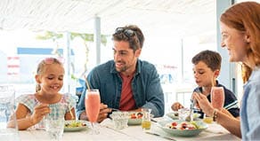 Cheerful family in restaurant enjoying lunch together in a patio  Mother and father observing daughter while eating food with son busy in eating  Smiling family with two children eating brunch together at kiosk during summer vacation 