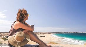 Female tourist looking the tropical panorama in a windy day  Amazing ocean beach in Fuerteventura Spain  Beautiful girl relaxing on a wooden bench  Travel, vacation, happiness and carefree concept