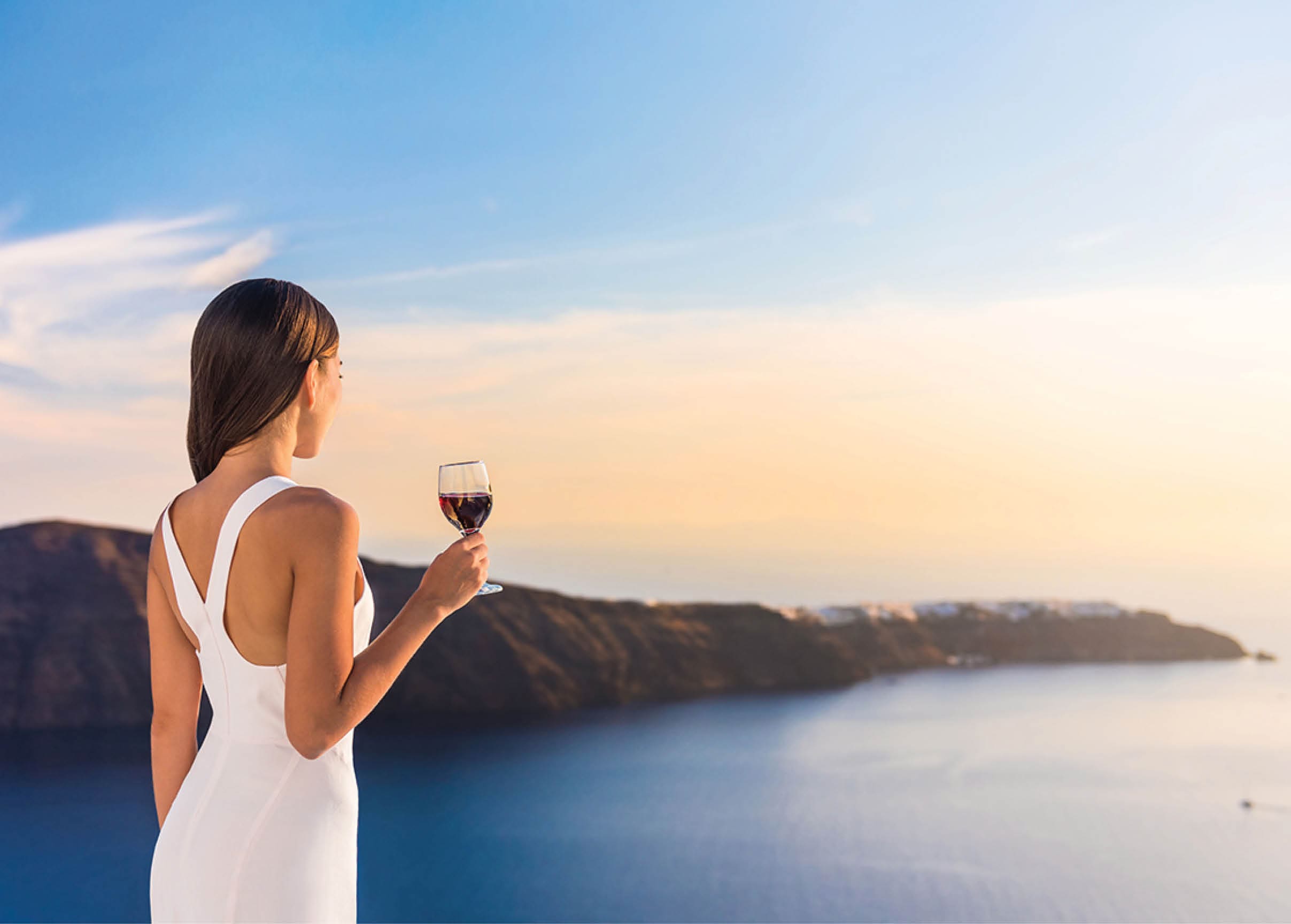 Young woman drinking red wine on outdoor terrace watching beautiful sunset view of Mediterranean Sea  Female in white sundress on summer Europe travel vacation in Santorini, Greece 