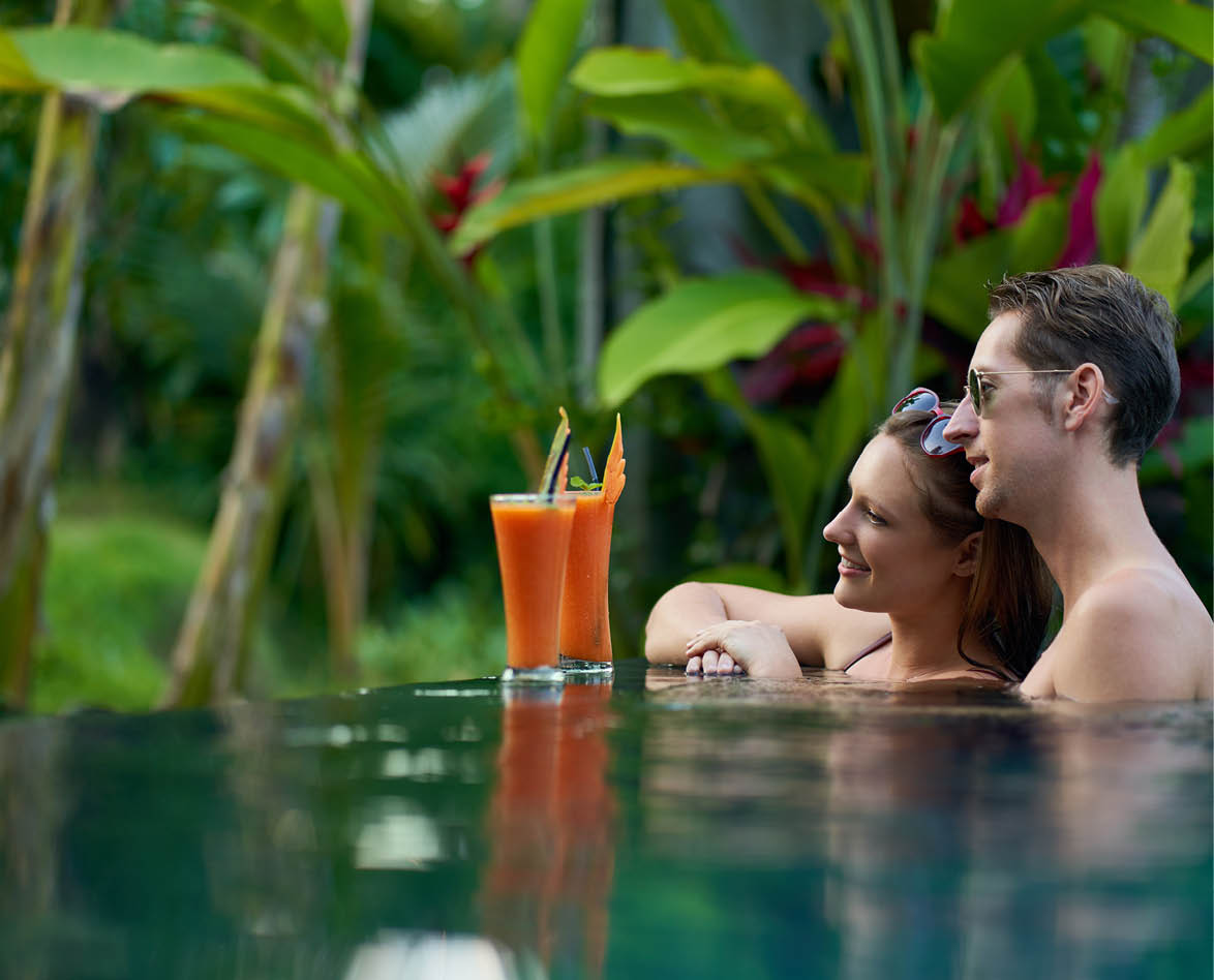 Happy millennial couple having cocktails together in pool of luxurious hotel and spa with tropical plants, wearing trendy bathing suits and sunglasses smiling