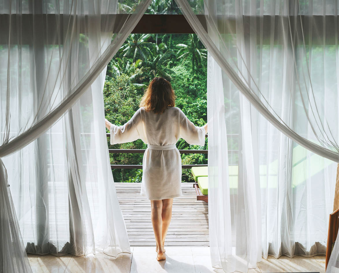 A woman in a bathrobe opens the curtains in deluxe Bali hotel room overlooking the terrace and tropical trees Woman is awoke and standing before window  Girl is opening curtains and meeting sunrise
