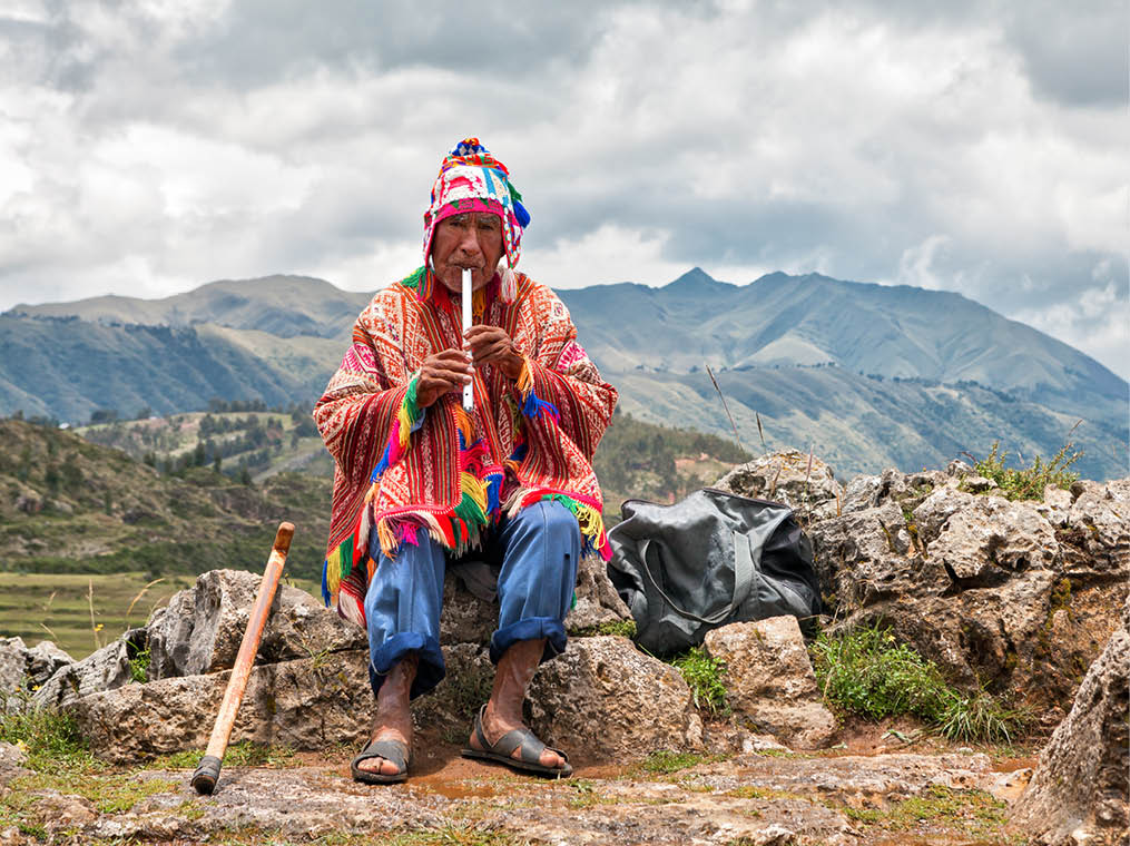 The old man Quechua dressed in a colored poncho and a cap of Chullo, sits on the rocks with a view of the mountains in the background and plays on the musical instrument of Quena  Cusco, Peru 