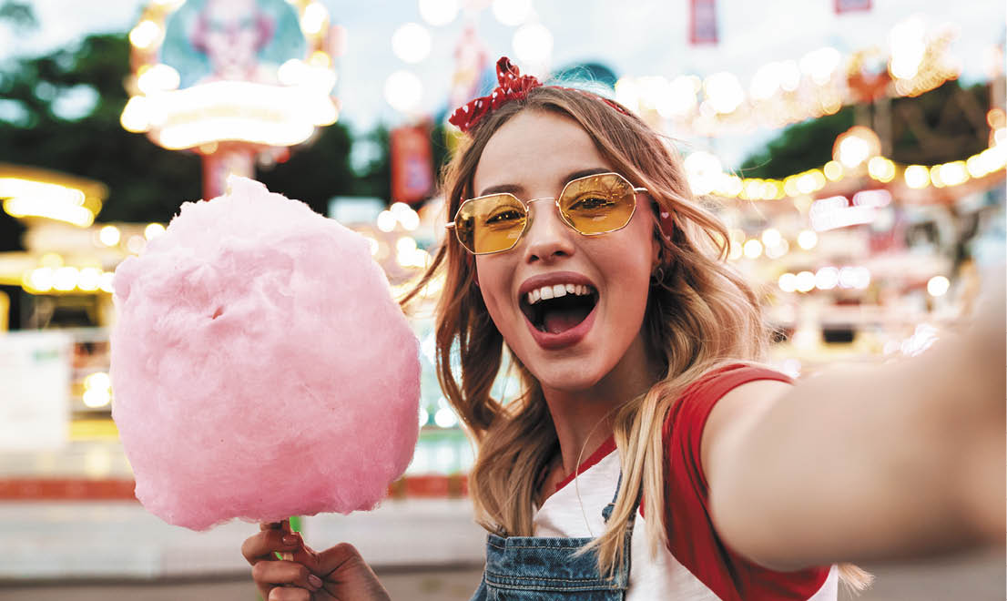 Image of excited blonde woman wearing girlish clothes holding sweet cotton candy while taking selfie photo at amusement park