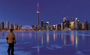 Man standing on frozen Lake Ontario watching Toronto city skyline light up at dusk